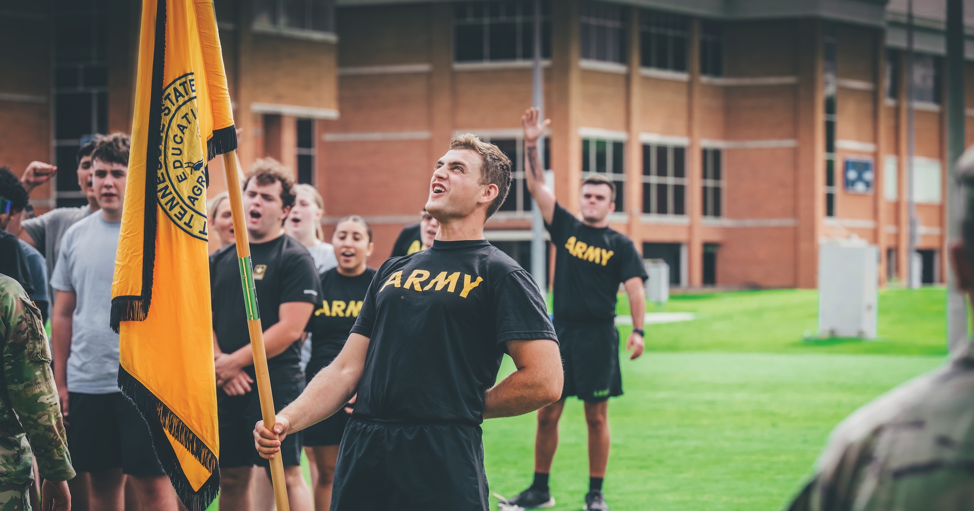 ETSU ROTC members wear Army t-shirts and participate in a drill outside. The person in front is holding a flag.