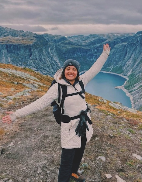 A Roan Scholar stands in front of a canyon with a lake, and her arms are outstretched.