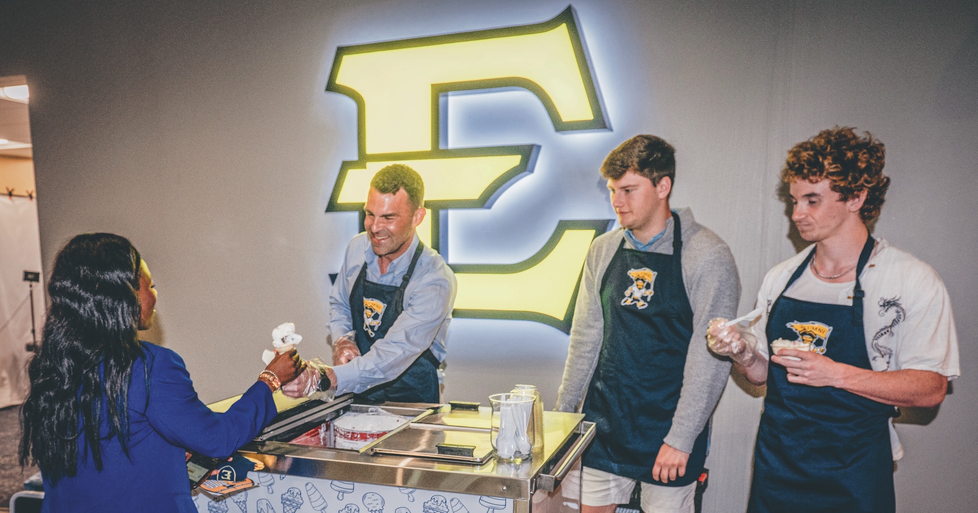 A person wearing a Forever ETSU apron hands an ice cream cone to a student during a Forever ETSU event in the Cave. Two other people wearing aprons and assisting look on.