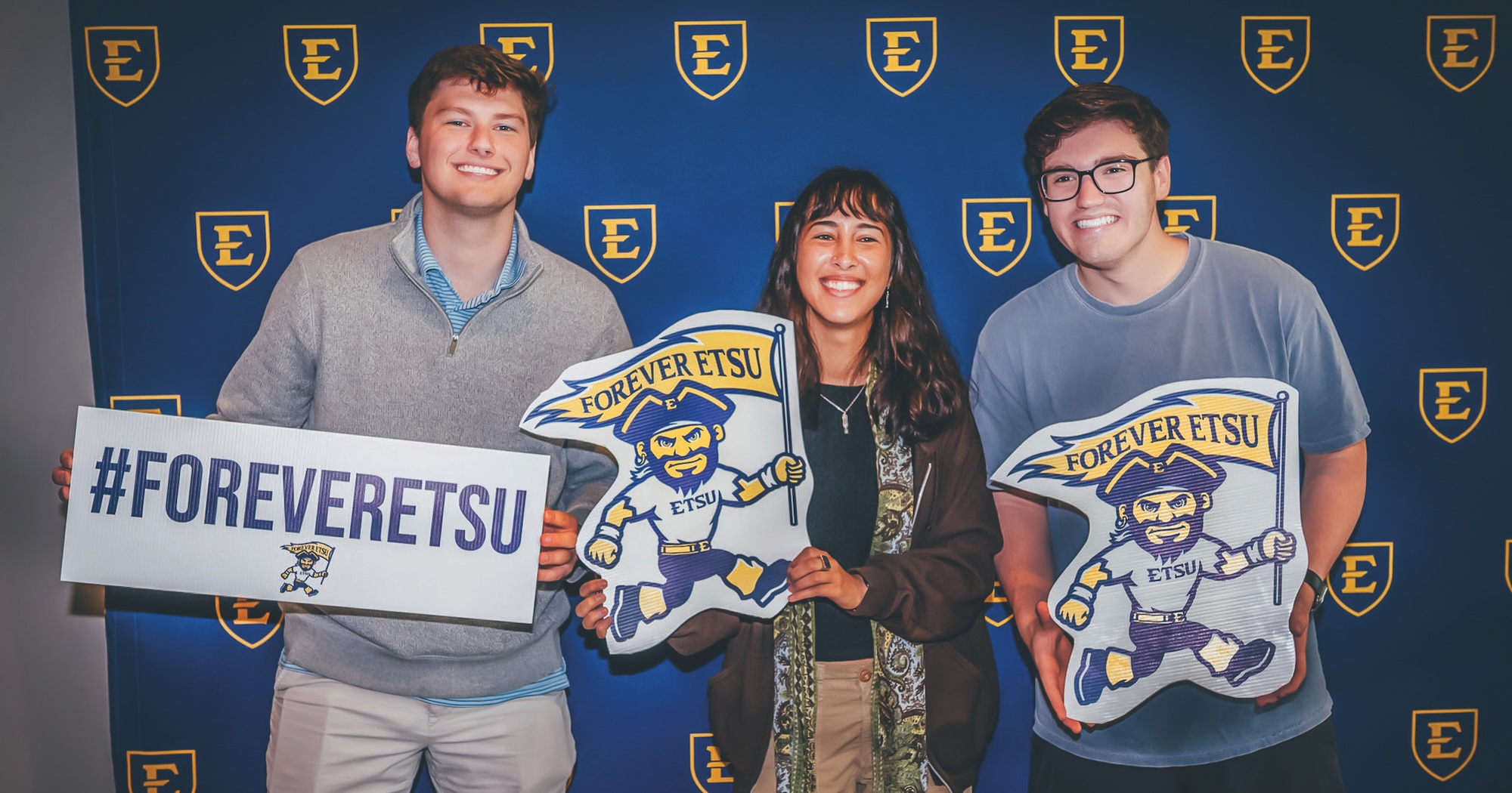 A person wearing a Forever ETSU apron hands an ice cream cone to a student during a Forever ETSU event in the Cave. Two other people wearing aprons and assisting look on.