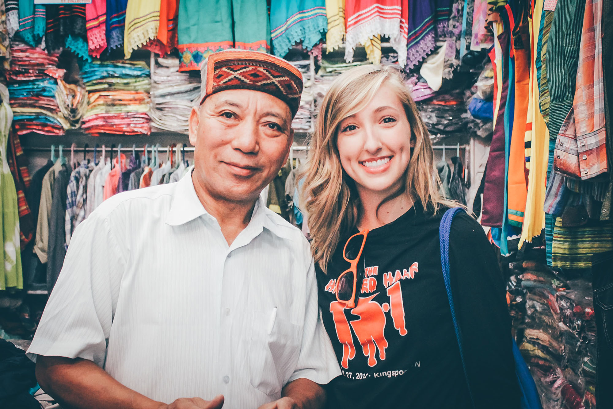 A Roan Scholar holds the poses with someone in their shop with brightly colored clothing and fabrics in the background.