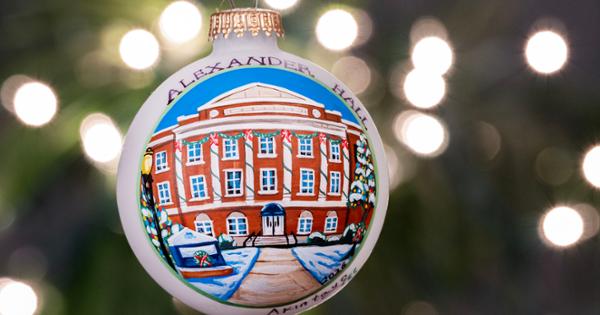 2026's holiday ornament, featuring Alexander Hall, in the foreground. Holoiday tree branches and lights light up the background.