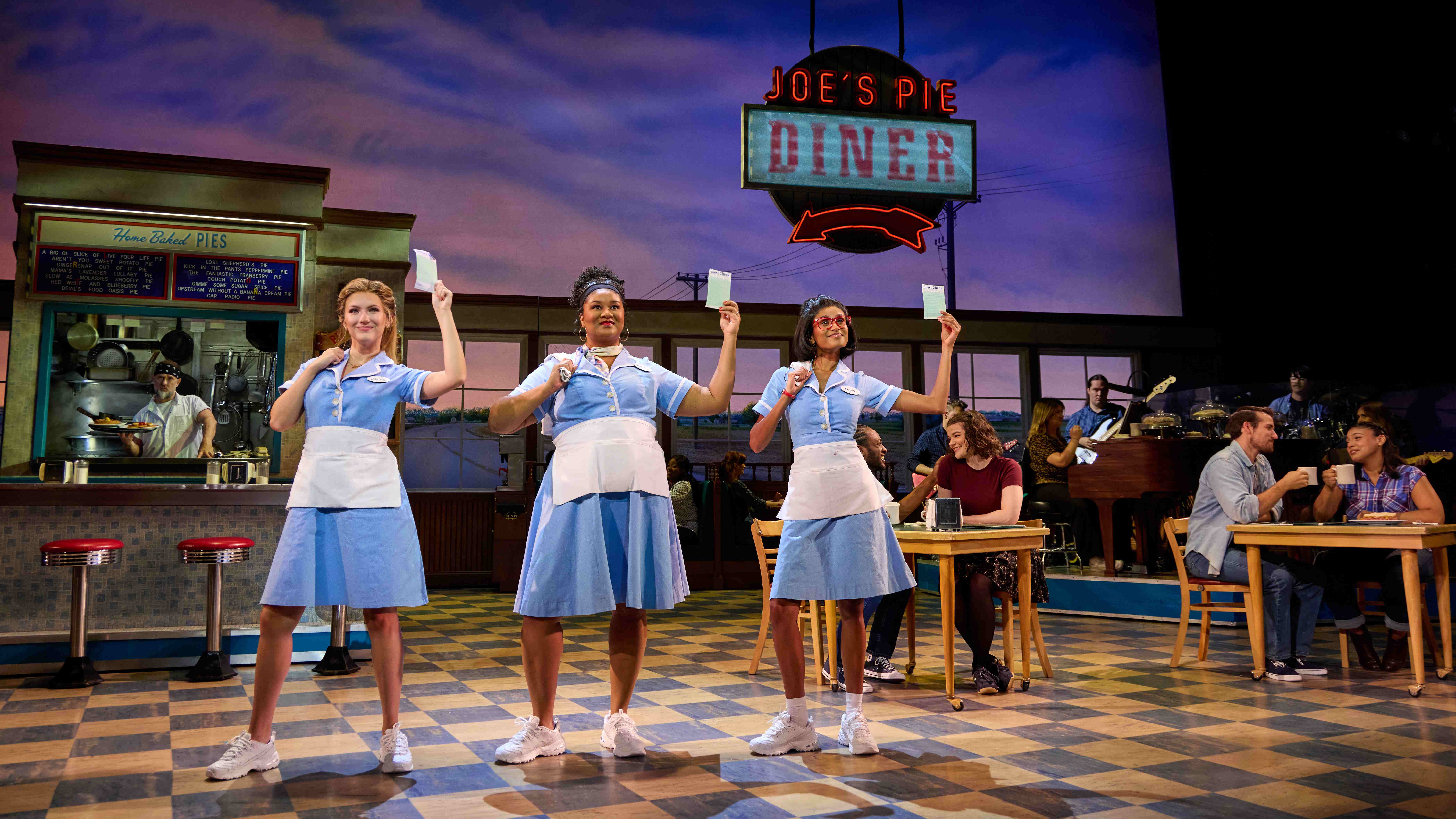 three waitresses in light blue diner uniforms holding up paper order tickets