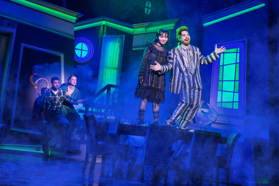 Stage photo from Beetlejuice featuring Lydia and Beetlejuice singing on a table, with two characters crouching nearby on a blue, moody set.