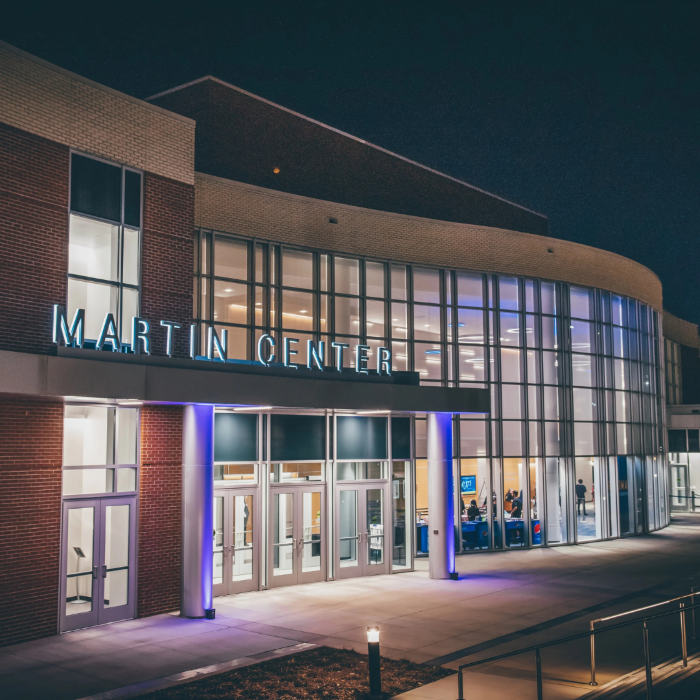 Nighttime view of the modern, glass-fronted Martin Center building entrance, lit up with blue lighting under a prominent sign reading "MARTIN CENTER".