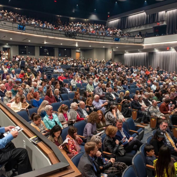 Perspective from the side balcony showing a large crowd of people filling the multi-tiered seating of a grand theater before an event.