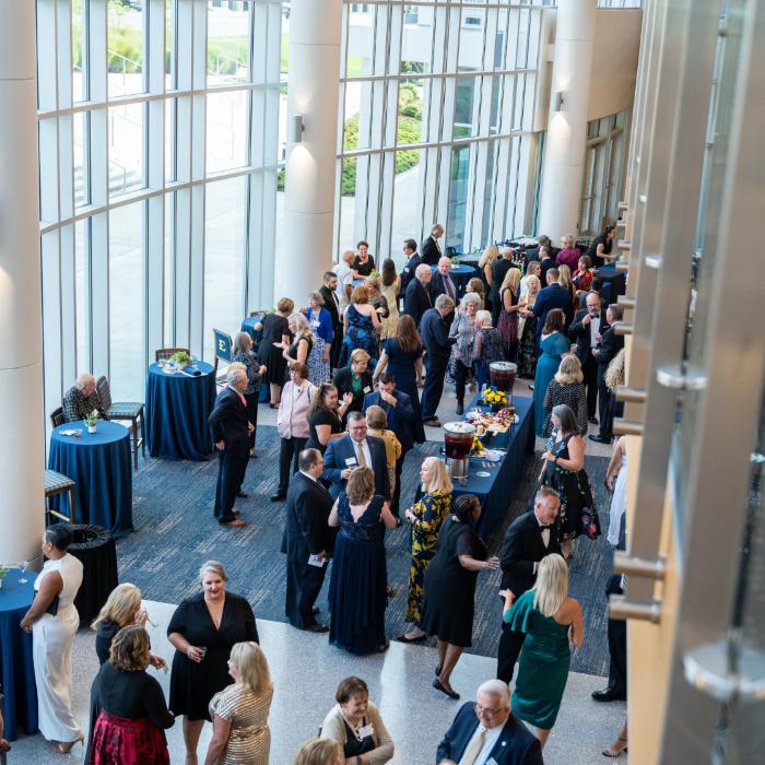 High-angle shot of a bright, spacious lobby with high glass walls where people in formal attire gather around drink tables during a reception.