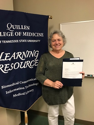Martha Whaley holds her official notice of promotion in front of a banner for the Department of Learning Resources