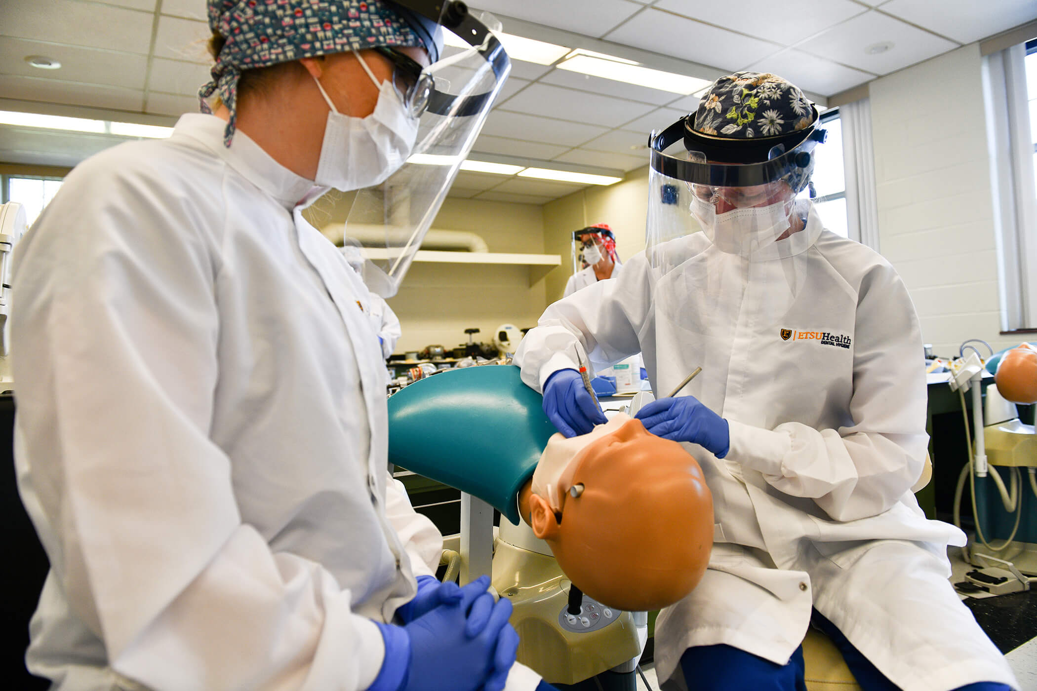 two students dressed in dental hygiene protective gear and lab coats perform skills exercises on a mannequin.