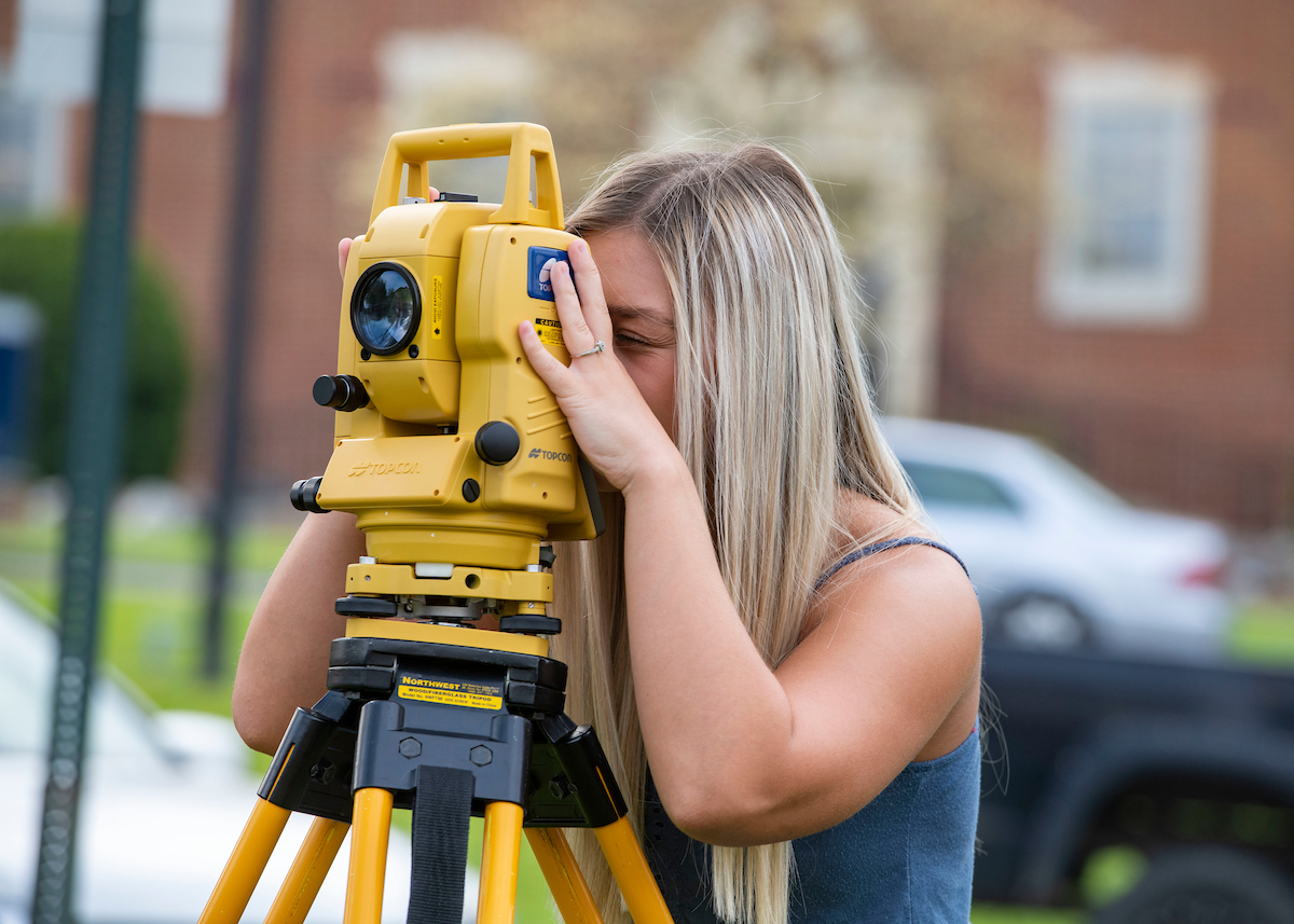 A college-aged woman looks through a piece of surveying equipment. She is on ETSU's campus.