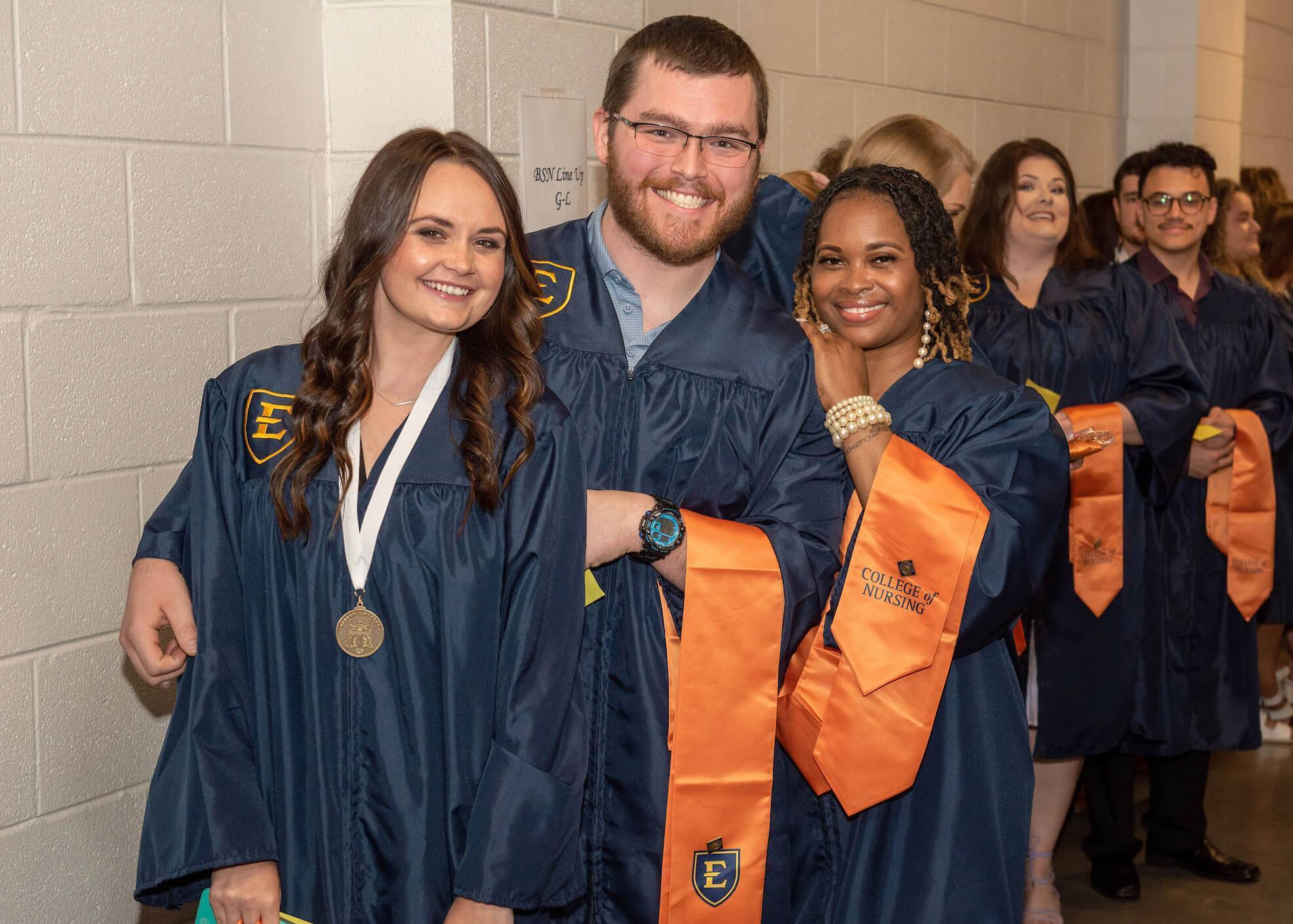 A line of students at graduation. Two women and a man smile at the camera.
