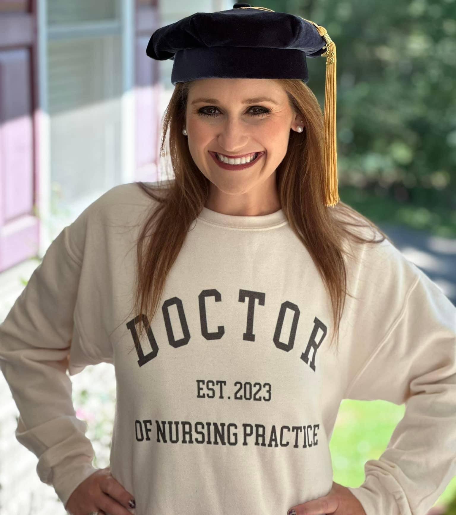 A woman stands outside, wearing a DNP sweatshirt and her doctoral hat.
