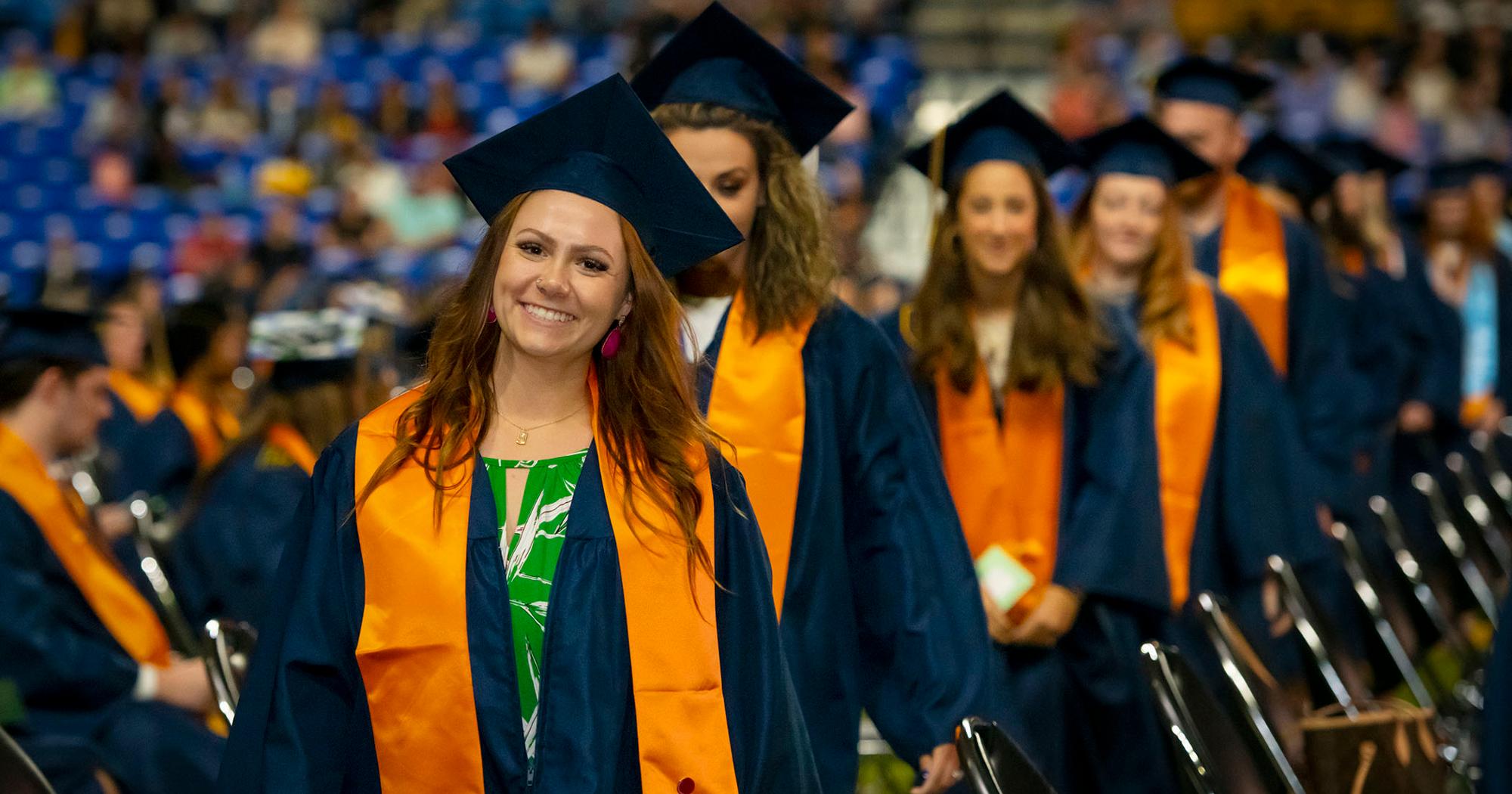 a woman smiles at graduation, in her cap and gown. She leads the way to the stage. She is the only one in focus.