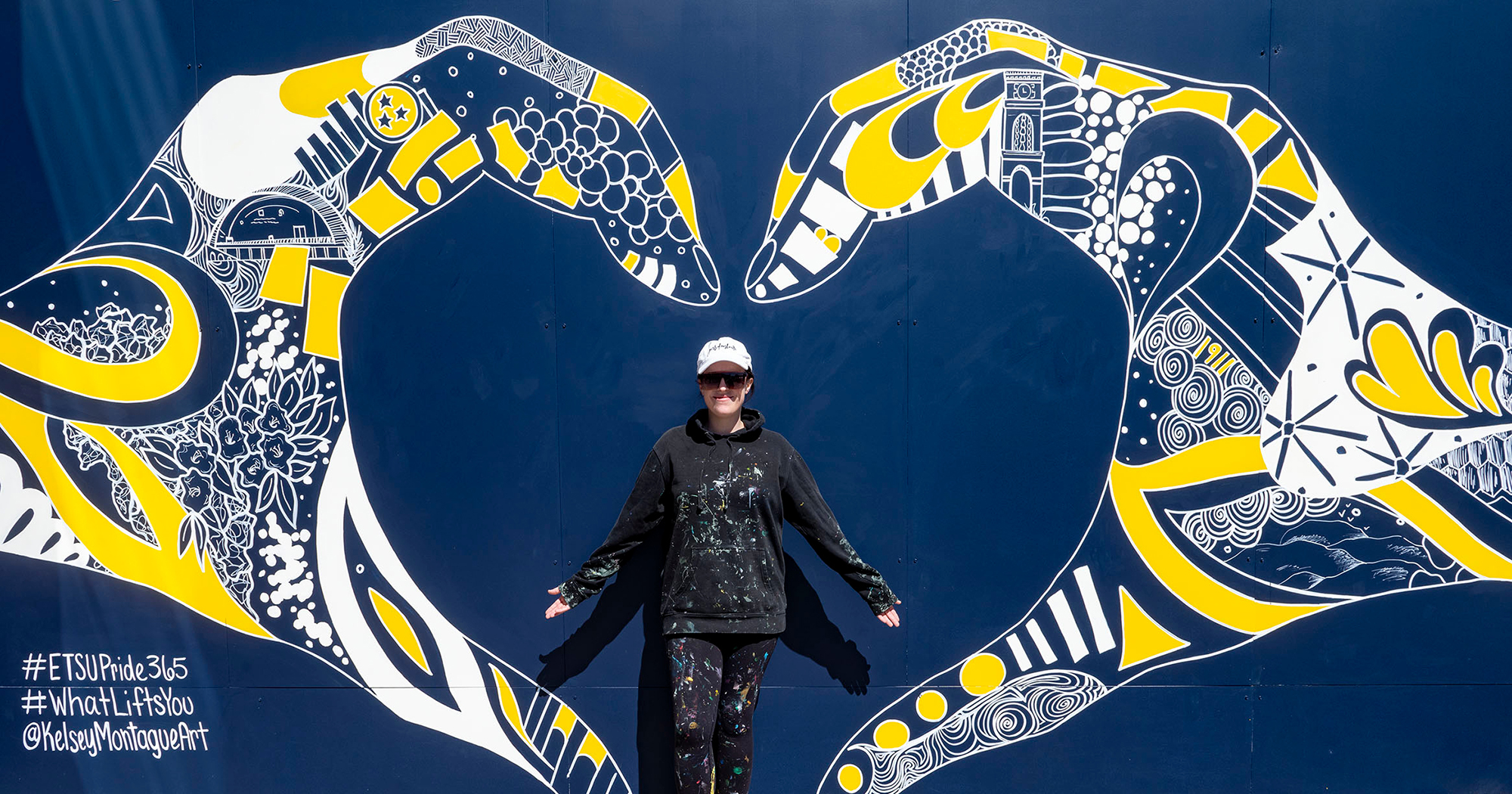 A mural of two hands joining together to create a heart. Inside the outlne of the hands are doodle-like drawings of regional symbols, such as rhododendrons and mountains as well as ETSU landmarks like the carillon. 