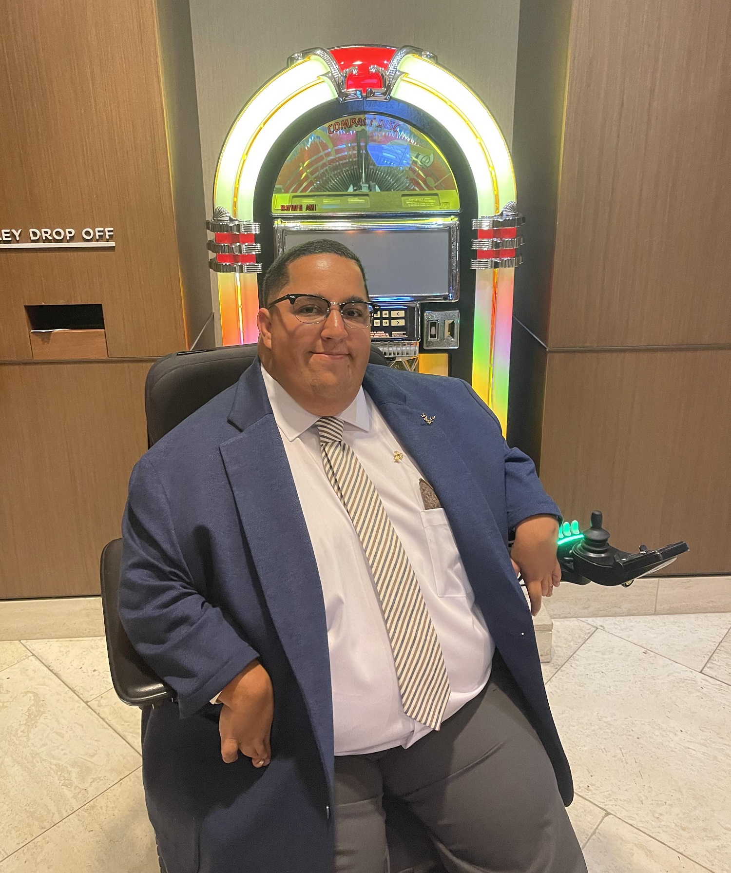 Greyson Jennings sits in his wheelchair in front of a colorful jukebox as he smiles for the camera. He is wearing a suit and tie. 