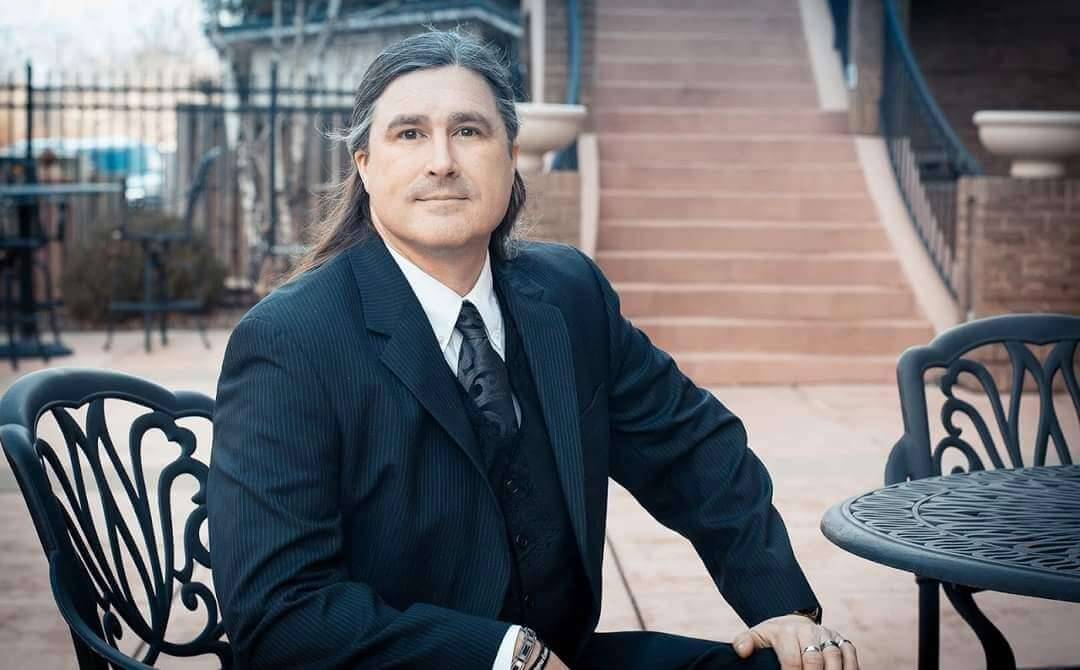 Man in a suit with long hair looks into the camera while sitting outside.