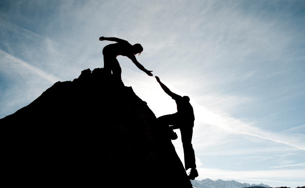 silhouette of one person reaching down to help another up a mountain